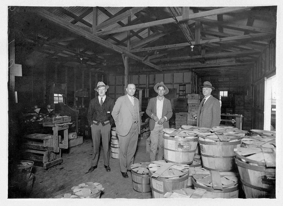 Four men standing among stacked bushel baskets and P&D crates inside the early Pittman & Davis packing house